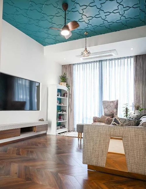 DESIGN CHRONICLES This living room, crafted by an interior designer in Mumbai, features a blue textured ceiling, a wooden herringbone floor, a wall-mounted TV, and cozy seating arrangements like an armchair and sofa.