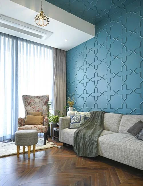 DESIGN CHRONICLES A modern living room designed by a luxury interior designer 7Nirvana features a textured blue accent wall, beige sofa, floral armchair, footstool, and wooden herringbone floor.