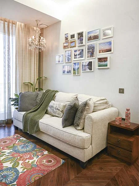 DESIGN CHRONICLES Cozy guest room with a beige sofa adorned with patterned cushions and a green throw, a colorful floral rug, wooden side table, and a gallery wall of framed photos under a crystal chandelier.
